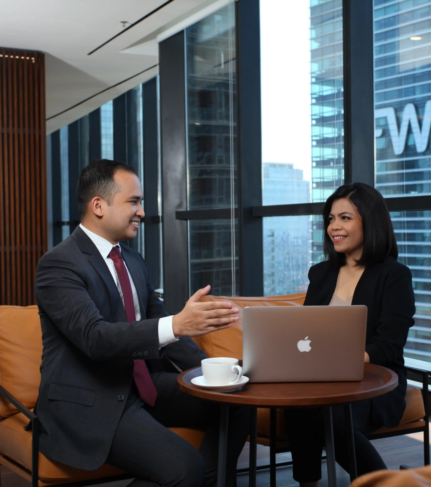 Man and woman in business attire sitting at a round table with a laptop and coffee, having a discussion in a modern office with glass windows
