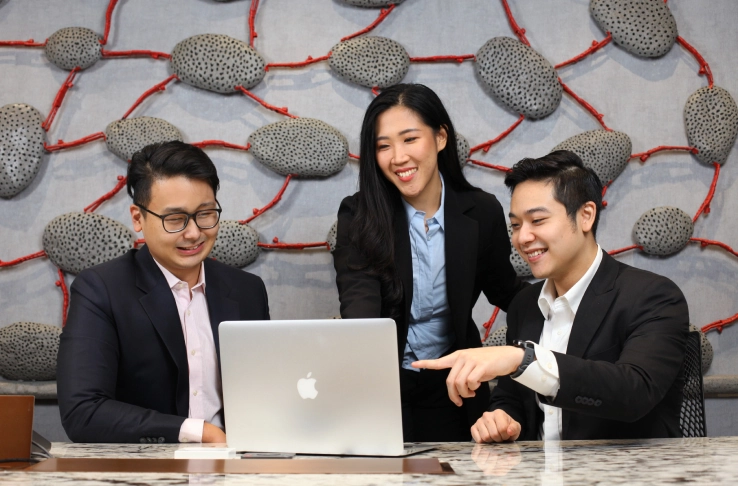 Three people in business attire gathered around a laptop, smiling and discussing, with a decorative stone wall background