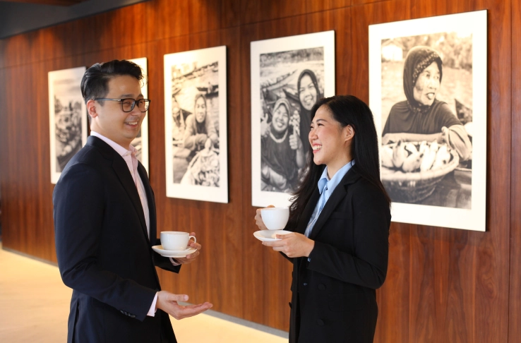 Man and woman in business attire holding coffee cups and talking in front of framed black and white photos on a wooden wall