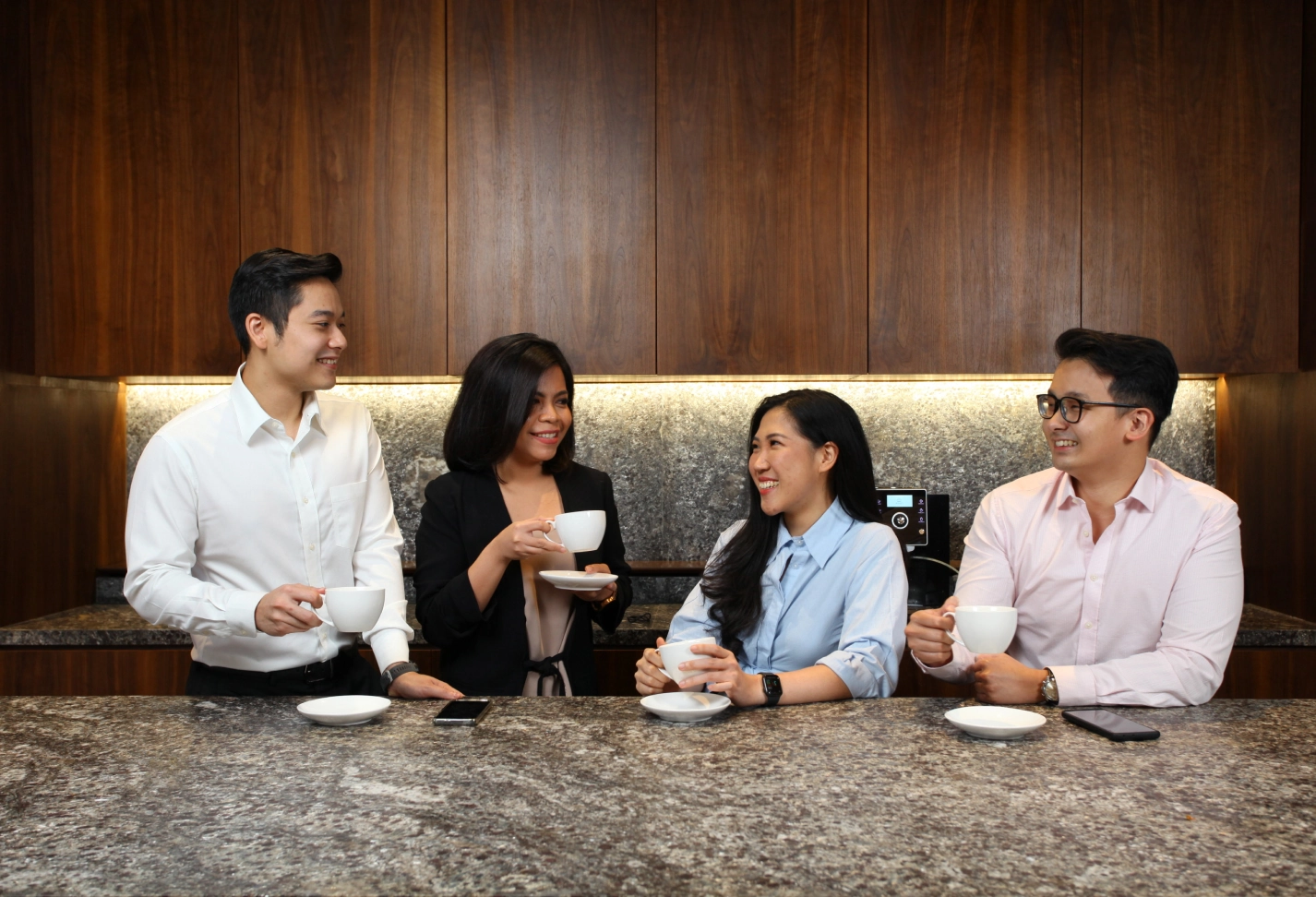 Four people in business casual attire standing and sitting at a granite counter, holding coffee cups and smiling, with a wooden wall background